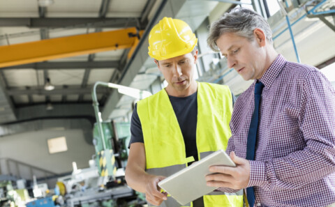 The Benefits of Outsourcing Health and Safety displayed by a worker and manager in a warehouse discussing plans over a tablet. The worker is wearing high visibility PPE and a hard hat.