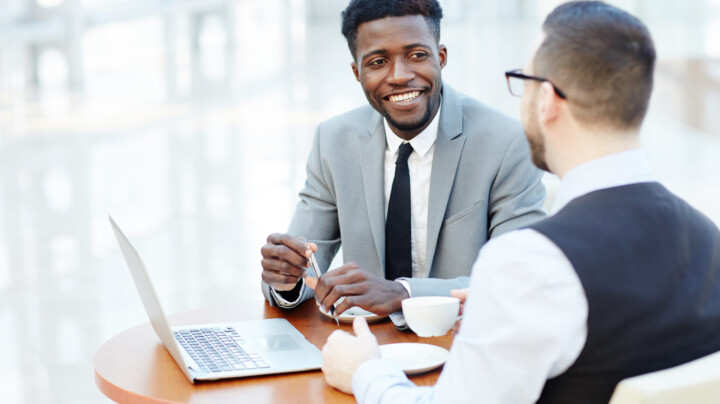 Portrait of successful African-American businessman smiling while discussing deal with Caucasian partner during meeting at coffee break to represent Benefits of Employee Wellness Programs article.
