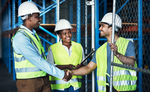 Impact of social values on business represented by three factory workers, two of which are shaking hands. They are dressed in high visibility vests and white hard hats.